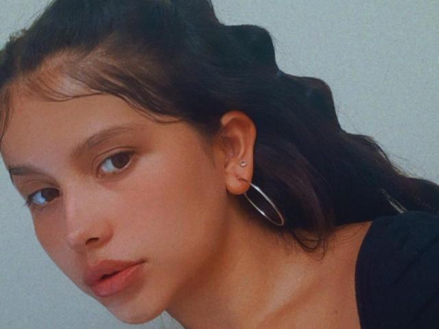 Melany White tiene un rostro adorable, con piel de color marrón claro, pequeños aretes redondos y un labio ligeramente entreabierto. Lleva una blusa negra y tiene el cabello oscuro y ondulado..