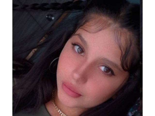 Melany White tiene el cabello largo y oscuro, ojos marrones y labios rosados, y lleva una blusa verde con una cadena dorada, mirando a la cámara con una sonrisa sutil..