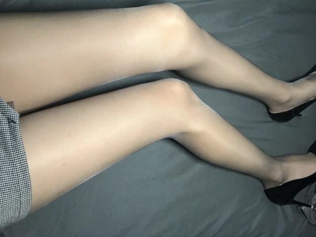 TinaFany ligt op een grijze bed, met aan haar benen dunne panty's, zwarte hoge hakken en een geruite rok. Haar bleke huid is glad, met subtiele lijntjes bij de knieën..