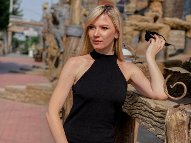 Paradise. Roxanne, con cabello rubio largo, vestido negro de tirantes, sosteniendo una tarjeta negra, apoyada en una estructura de madera, sonriendo, piel clara, delgada, senos pequeños, iluminada por el sol, en un entorno al aire libre..