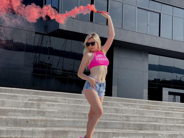 ParadiseRoxanne mit blonden Haaren, einem pinken Crop-Top und Jeansshorts, die roten Rauch aus einer Pfeife pustet, trägt schwarze Sonnenbrillen und steht auf einer Betontreppe. Die Brüste sind sichtbar..