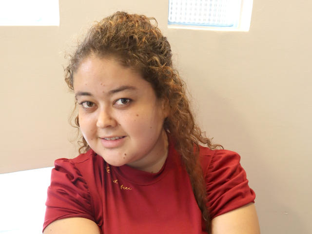VanessaRosse, avec ses cheveux bruns bouclés, son t-shirt rouge, son léger sourire et ses petites taches de rousseur sur le visage, assise les bras croisés, regardant la caméra..