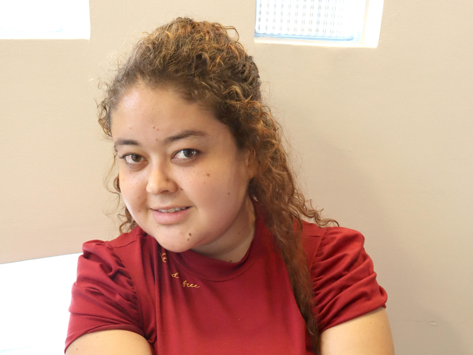 VanessaRosse with curly brown hair, red shirt, slight smile, and small moles on her face, sitting with arms crossed, looking at camera.