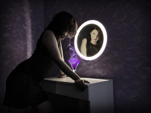 AmethystCharm with curly dark hair, black lace dress, leaning on white vanity, staring at circular mirror, purple magic glow, illuminated face, sexy, mysterious, dark room.