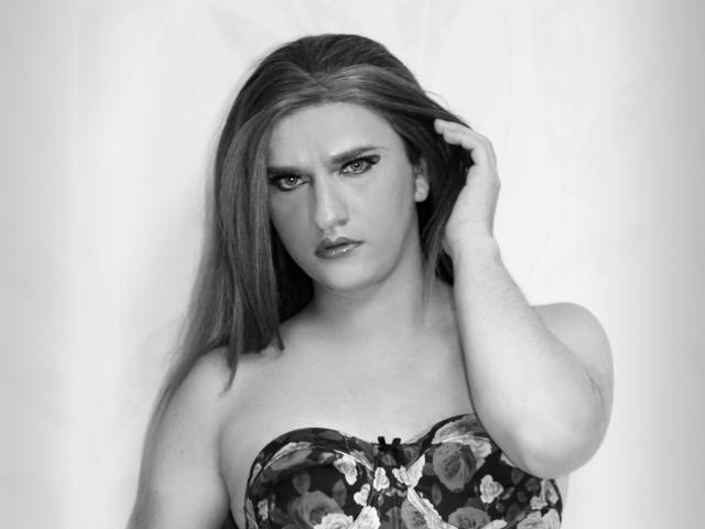 Black and white photo of TriniTs with long, straight hair, gazing intensely, hand in hair, wearing a floral strapless top, showing her tits, and a serious expression.