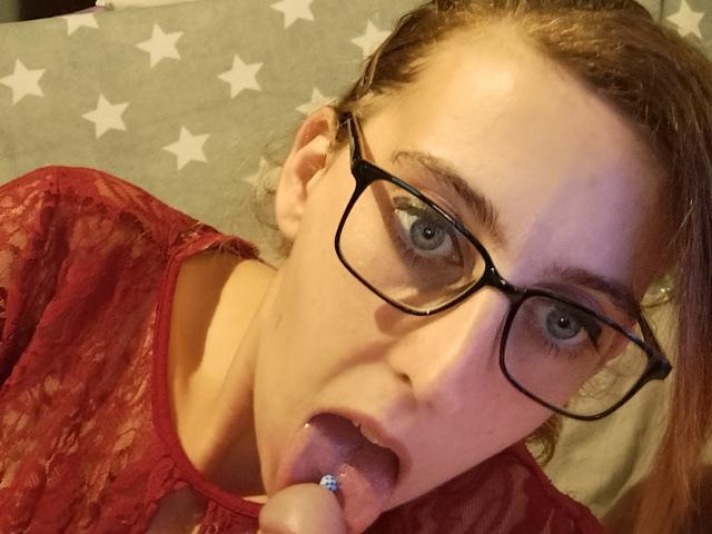 Coquine met een bril, rode kanten top, tong uit, zuigt aan een doorboorde vinger, blauw oog, lichte huid, bruin haar, lichtelijk rommelig, kijkt omhoog, mond open, zachte belichting..