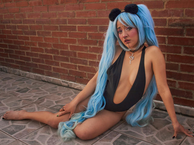 SalomePrince with long blue hair, black pom-pom headband, and deep black one-piece showing tits and large ass, sitting on tiled floor, wearing key necklace, against brick wall.