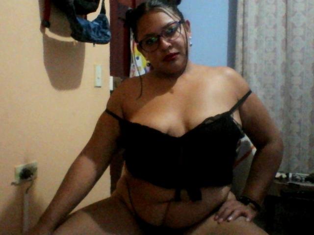 Adi69, avec des lunettes, un soutien-gorge en dentelle noire et des sous-vêtements noirs, assise les jambes écartées, montrant ses grandes seins et son ventre, les cheveux foncés attachés en chignons, rouge à lèvres, et un sourire confiant..