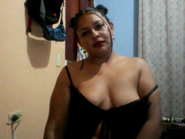 Adi69 with dark hair in buns, wearing a black, low-cut top, exposing her big tits, red lipstick, and earrings, standing in a room with a wall and curtain.