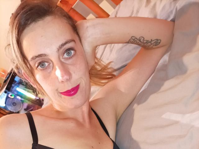 Eine Frau mit blauen Augen, rotem Lippenstift, einem schwarzen BH, einem Armtattoo, die auf einem Bett liegt, lächelt, hat blondes Haar, eine Hand hinter dem Kopf, ein weißes Bettlaken, ein hölzernes Kopfteil, ein sichtbarer Fernseher im Hintergrund..