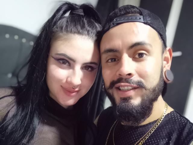 SativaAndGaspar69, con capelli neri e ombretto viola, con un anello al naso, sorride a Gaspar69, che ha barba, baffi e un berretto nero, indossa una camicia a pois e una catena d'oro..