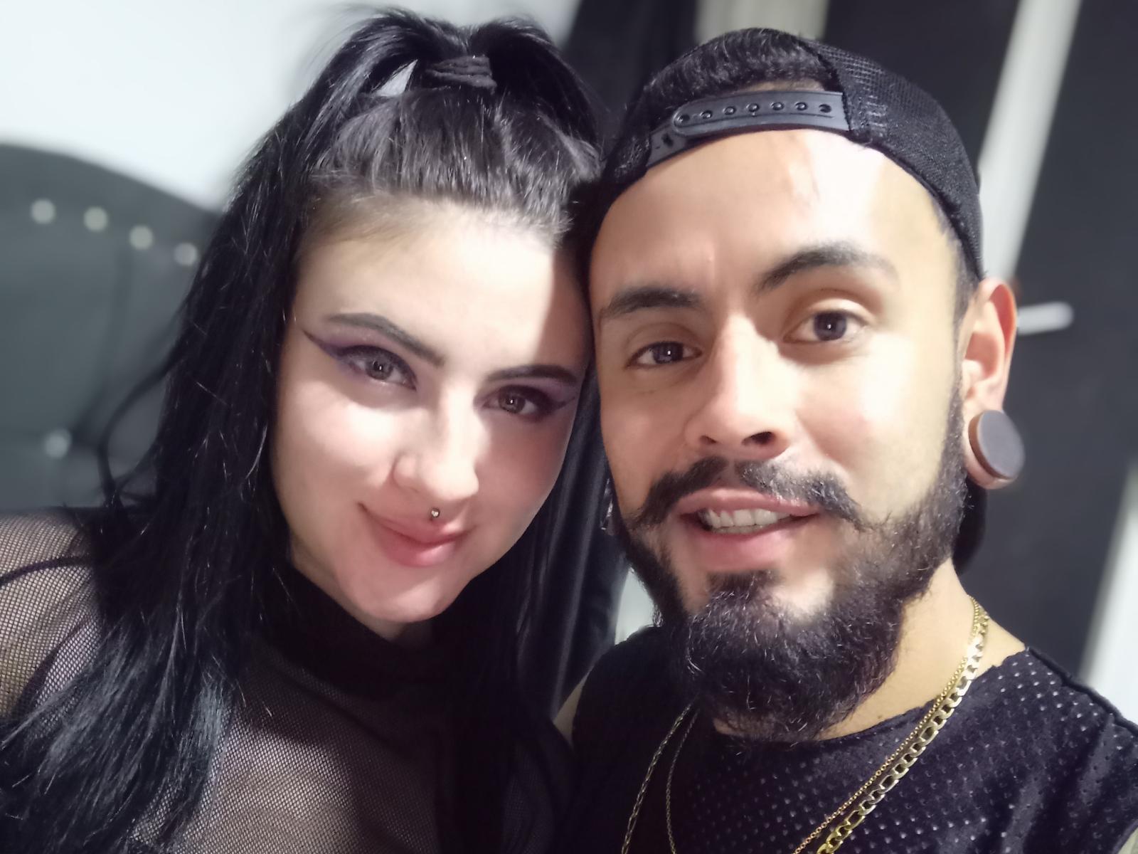 SativaAndGaspar69 mit schwarzen Haaren und lila Lidschatten, Nasenring, lächelt Gaspar69 an, der einen Bart, einen Schnurrbart und eine schwarze Mütze trägt, ein gepunktetes Hemd und eine goldene Kette..