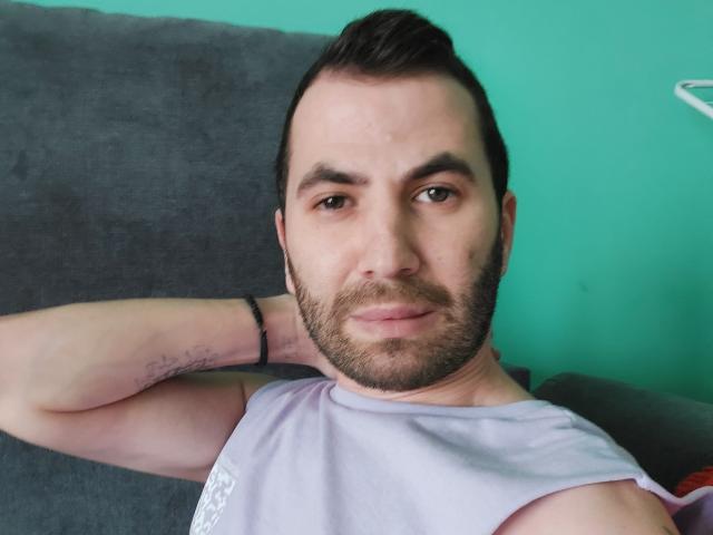EnddyHottx, mit leichtem Bart, kurzen, dunklen Haaren und einem Tattoo auf seinem rechten Arm, trägt ein violettes, ärmelloses Hemd und hat einen entspannten, leicht selbstgefälligen Gesichtsausdruck..