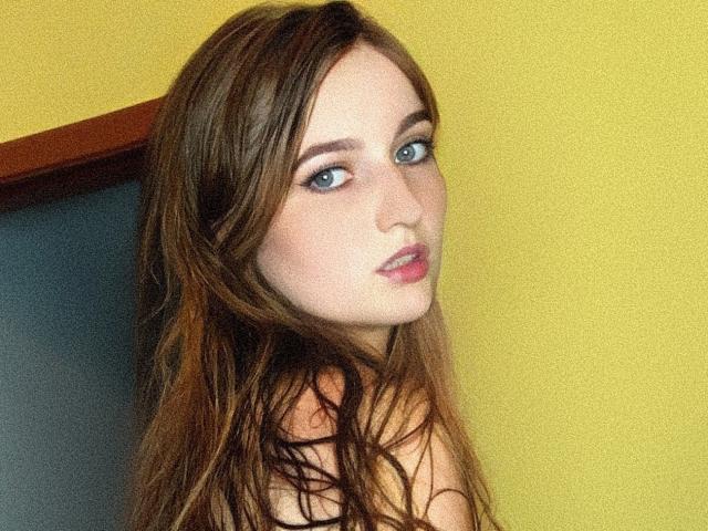 EmmyRay, com cabelos castanhos longos, olhos azuis e lábios rosados, olhando por cima do ombro com um leve franzir, mostrando seus ombros nus contra uma parede amarela..