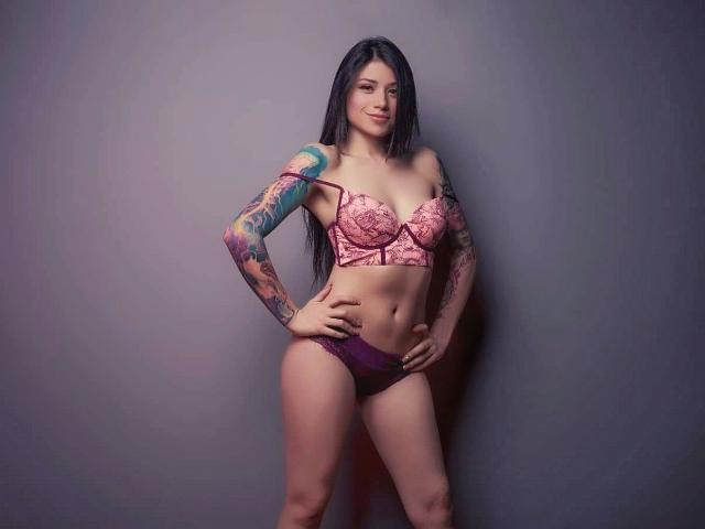 Alice Leblanc, con lunghi capelli neri, reggiseno di pizzo rosa e mutandine viola, braccio tatuato, in piedi con le mani sui fianchi, sorridente, che mostra il seno e i glutei, in uno studio fotografico..
