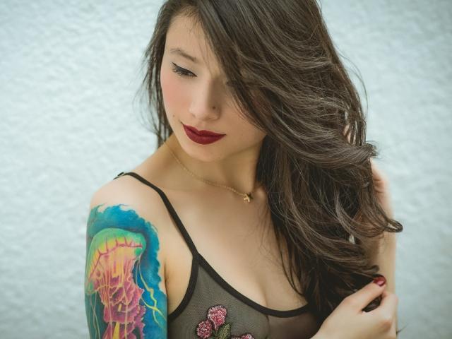 AliceLeblanc, com cabelos castanhos longos, batom vermelho escuro, blusa de malha preta, tatuagem colorida no braço direito, segurando o cabelo, expressão suave, pele clara, colar de ouro pequeno, em pé..