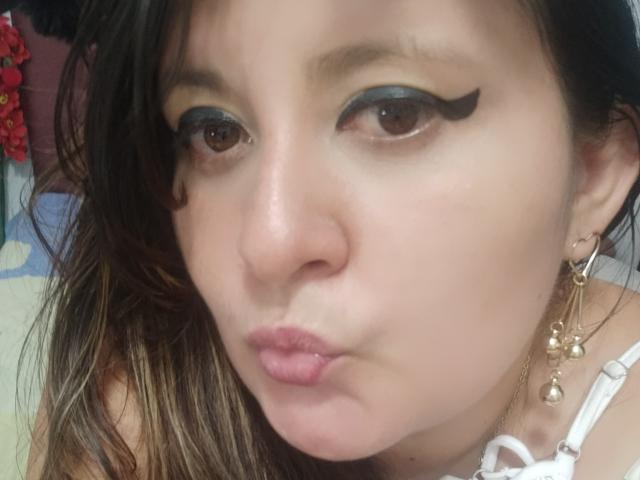HotValeryPassione, met volmaakte lippen, draagt zwarte eyeliner, gouden oorbellen en een witte jurk met bandjes. Ze heeft bruine ogen, golvend bruin haar en een lichte huidskleur..