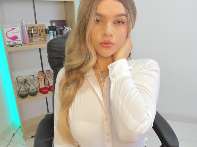 ValerieStone with long blonde hair, pouty pink lips, white button-up shirt, hand on cheek, necklace, sitting in chair, shelf with makeup and red heels in background.