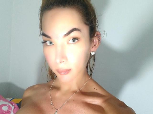 BarkiEscarleth, con piel morena, cabello castaño recogido en una coleta, ojos azules, cejas arqueadas, nariz pequeña, maquillaje discreto, pequeños aretes, collar y un hombro descubierto, mirando a la cámara..