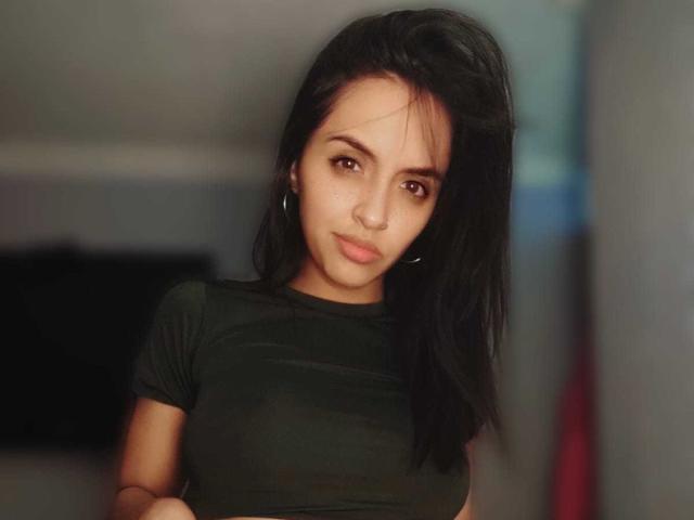 Chelsee with black hair, green eyes, and small nose, wearing a tight green shirt, looking at the camera with a slight smile, showing her small tits and flat ass.