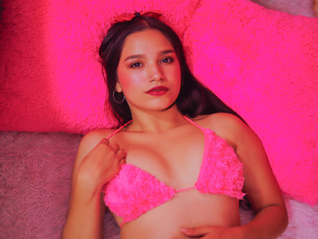 Demar, avec de longs cheveux noirs, un soutien-gorge rose duveteux, tenant les bretelles, des boucles d'oreilles cercles roses, allongée sur des coussins roses, éclairage rose, expression douce, main sur le ventre, rouge à lèvres rose..