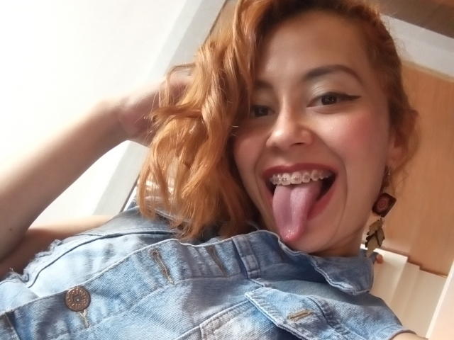 Eliza Jones tem cabelo ruivo, um colete de jeans azul e está de língua para fora, com aparelho, sorrindo para a câmera. Sua mão está no cabelo, mostrando os brincos..