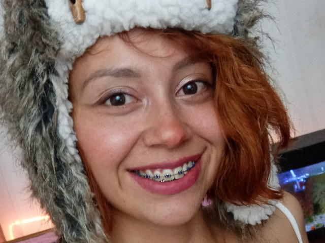 Eliza Jones, con cabello rojo rizado, usando un sombrero de lobo esponjoso de color gris y blanco, sonriendo y con brackets metálicos en los dientes, lápiz labial rosa y aretes rosas..