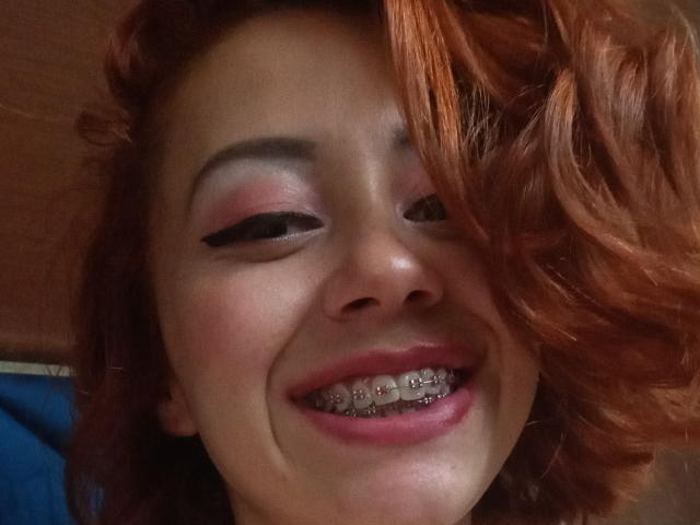 ElizaJones with red curly hair, pink eyeshadow, black eyeliner, and metal braces, smiling with her pink lips, showing her teeth. Her nose is slightly upturned.