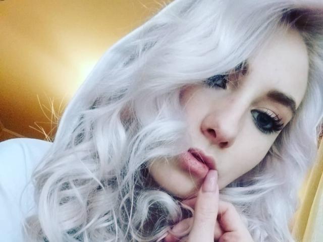 EllenSoNice, com cabelo branco e encaracolado, olhos azuis e lábios rosados, segurando o dedo na boca, com uma expressão sedutora. Seus cílios são longos e sua pele é clara..