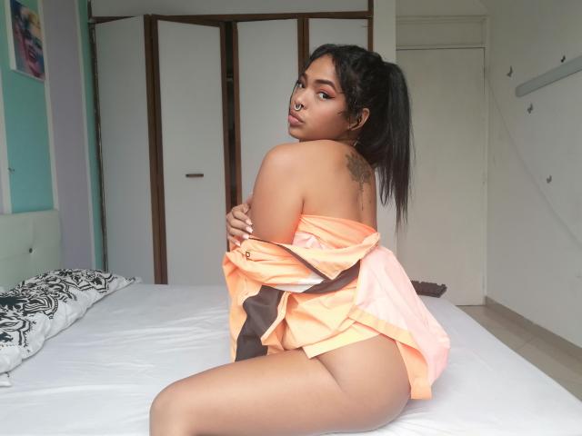 VickyGreys with tan skin, black ponytail, and tattoo on her back, sits on a bed, wearing an orange shirt, exposing her ass, looking over her shoulder, arms crossed.