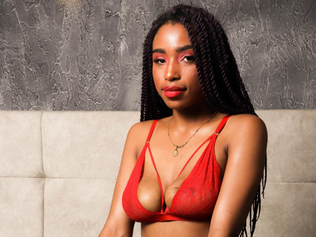 SiaHotty, peau foncée, portant un soutien-gorge en dentelle rouge et des cheveux tressés, montrant ses petites seins, avec du fard à paupières rose et du rouge à lèvres rouge, assise sur un canapé beige..