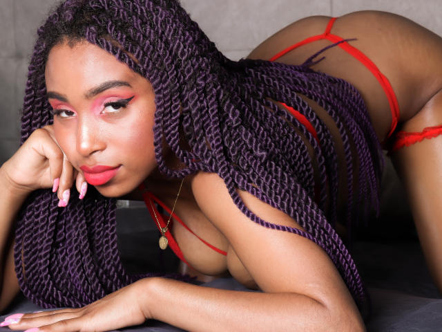 SiaHotty mit lila Zöpfen, roter Unterwäsche und pinkem Augen-Make-up, auf allen Vieren, zeigt ihren Hintern und den roten String, mit einem verführerischen Ausdruck und pinken Nägeln..