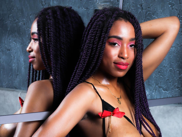 SiaHotty com tranças roxas escuras, sombra rosa e batom vermelho, vestindo um sutiã preto e vermelho, braço levantado, reflexo no espelho, pele brilhante, expressão confiante, colar de ouro pequeno..