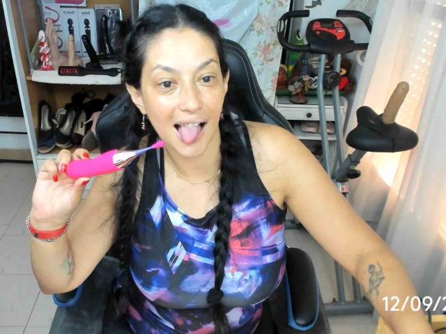 WetSweetKim con trenzas negras, lengua afuera, sosteniendo un vibrador rosa, usando un top deportivo con estampado de galaxia, con tatuajes visibles, sentada en una silla, sosteniendo una cámara para tomarse un selfie, fondo de cortina blanca..