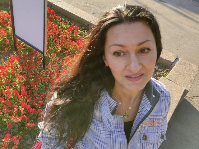 Antralyn with long dark hair, wearing a plaid shirt, necklace, and earrings, standing next to bright red flowers, looking up at the camera with a slight smile.