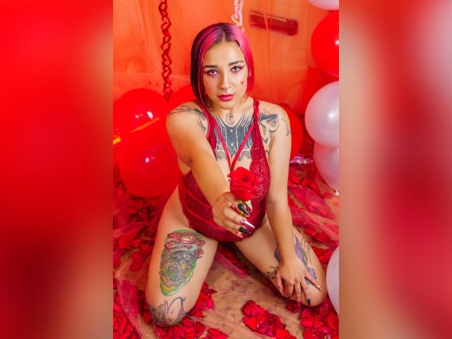 Lara Carter mit rosa und roten Haaren, Tätowierungen an Armen und Oberschenkel, rotem Spitzendessous, kniet auf roten Blütenblättern, hält eine Rose, rote Luftballons und weiße Luftballons, verführerischer Blick..