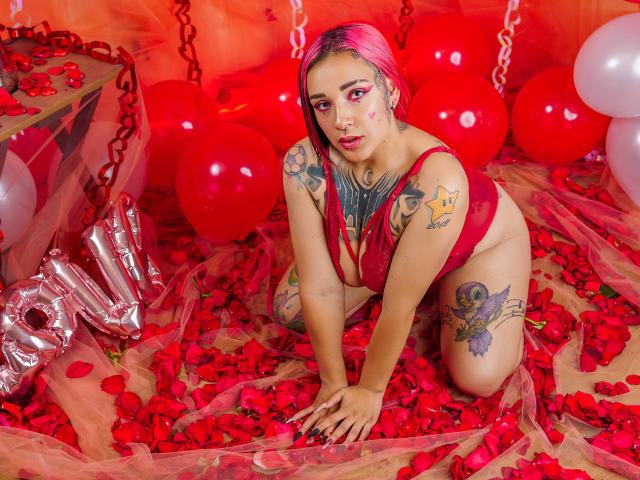 Lara Carter, mit rosafarbenem Haar, kniet auf roten Rosenblättern und trägt einen roten Spitzen-BH. Ihre Brüste und Tattoos sind sichtbar. Sie ist umgeben von Ballons und metallischen Zahlen. 1.
