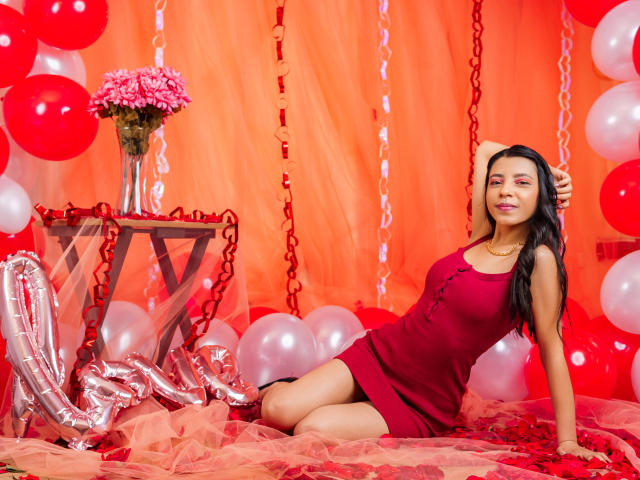 VannesaFlory in a red dress, sitting on a pink tulle, surrounded by red and white balloons, with a vase of pink flowers on a silver table. She's smiling.