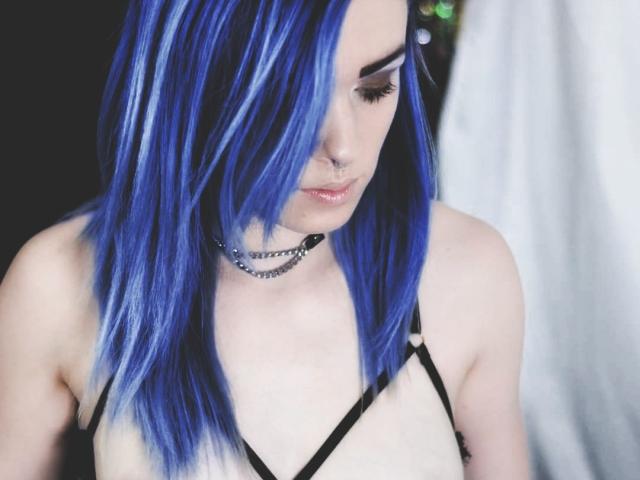 DreamArinna con capelli a righe blu e bianche, collana nera, piercing al naso, che guarda in basso, indossa un top con spalline nere, che mostra il seno, ha la pelle pallida e un'espressione sottile e sensuale..