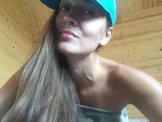 LilyOCute, con cabello castaño largo, usando un gorro verde azulado y una camiseta de camuflaje, mostrando sus senos y mirando a la cámara con una ligera sonrisa, en una habitación de madera..