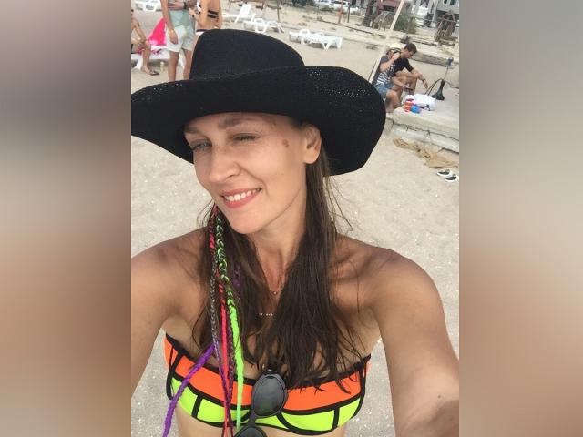 LilyOCute, com longos cabelos castanhos, usando um chapéu de cowboy preto e um top de biquíni laranja neon, sorrindo para a câmera, mostrando seios pequenos e tranças coloridas em uma selfie na praia..