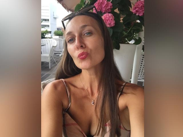 Lily, com lábios rosados e expressivos, cabelo castanho liso, olhos azuis, seios pequenos, vestindo um vestido com alças finas, um colar e óculos de sol na cabeça, sentada ao ar livre com flores rosa..