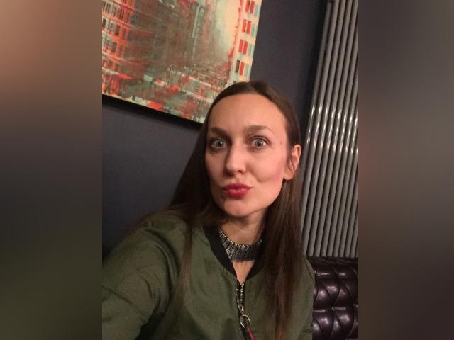 Lily, com cabelo castanho liso, camisa verde, colar metálico e batom rosa, fazendo beicinho, olhos azuis e um nariz pequeno, em um quarto com pouca luz..