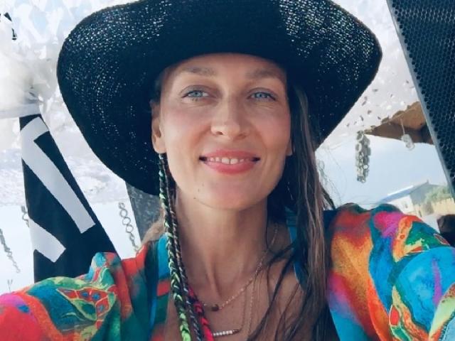 Lily, com olhos azuis, usando um chapéu preto, uma camisa florida colorida e cabelo trançado verde e rosa, sorrindo. Seus seios estão ligeiramente visíveis sob a camisa..