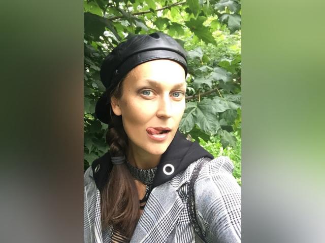 LilyOCute tiene ojos verdes, una coleta marrón trenzada y un gorro de cuero negro. Está sacando la lengua y lleva una chaqueta gris a cuadros. Su trasero no es visible..