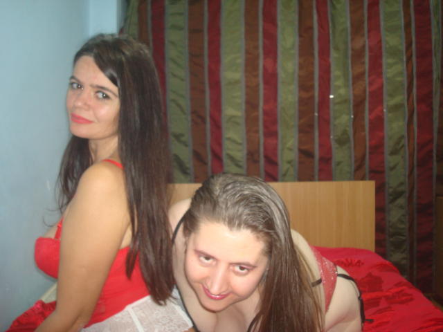 Two Sexduox women with long brown hair, one in red lingerie, the other in black, on a red bed, smiling at the camera, with the redhead in front.