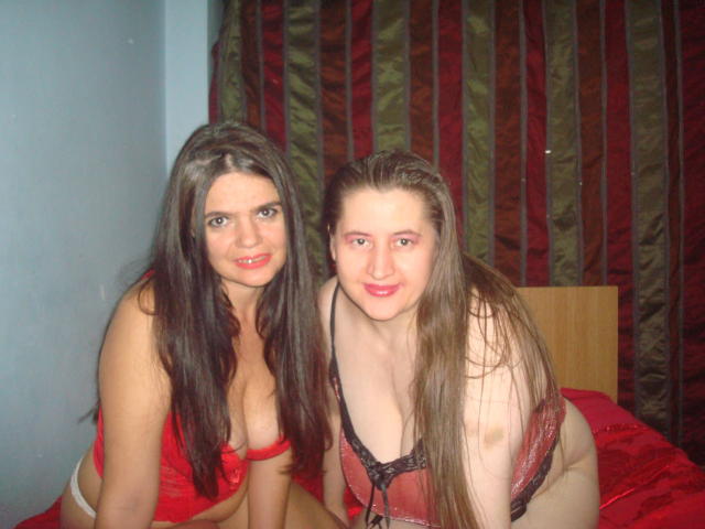 Zwei Frauen mit langen, braunen Haaren, rotem Lippenstift und freizügiger Unterwäsche, sitzen auf einem Bett. Eine hat große Brüste in Rot, die andere hat kleine Brüste in Schwarz..