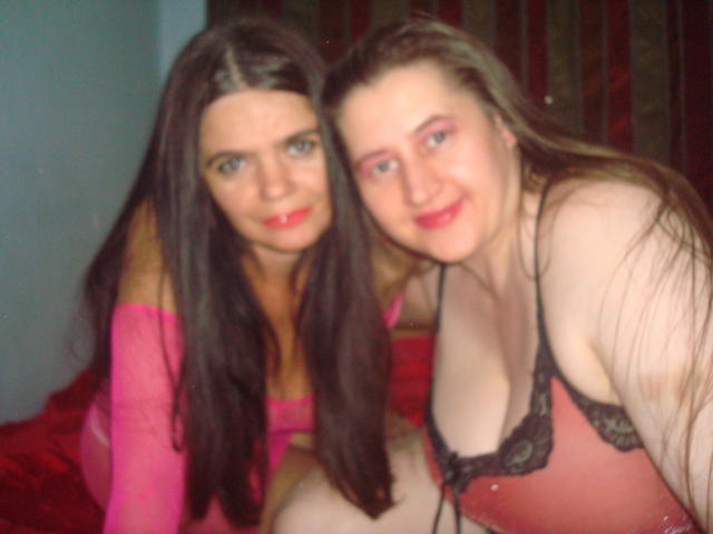Two Sexduox with long dark brown hair, pink off-shoulder tops, and red lipstick, sitting on a red bed, smiling at the camera, showing their tits in black lace bras.