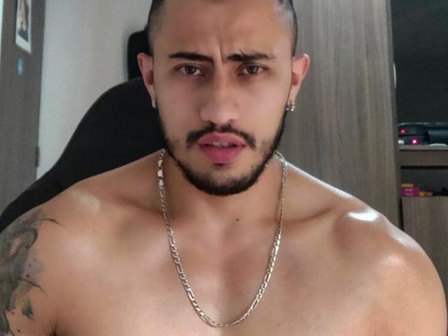 DimitriRivas sin camisa, con un collar de cadena, un tatuaje en el hombro derecho, perforaciones en las orejas y una ligera expresión de ceño fruncido. Su piel está bronceada y tiene una barba recortada..