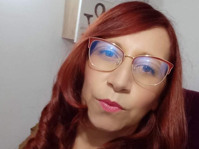 ValeryySweet, met rood haar, die gouden brillen draagt, een uitstekende roze mond heeft en blauwe, reflecterende ogen, ziet er een beetje grappig uit. Haar borsten zijn niet zichtbaar, alleen haar gezicht is te zien..